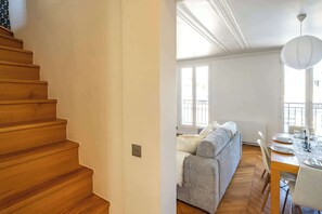 Una escalera de madera conduce a una sala de estar con sofá, mesa de comedor y vistas al exterior.