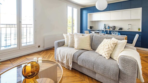 A living room with a grey sofa, white cushions, a glass coffee table, and a blue kitchen in the background. - 6 Guests Duplex - Martyrs Village with view (Paris)