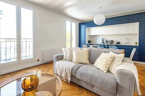 Un soggiorno con un divano grigio, cuscini bianchi, un tavolino in vetro e una cucina blu sullo sfondo.