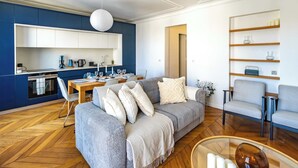 A modern living room with a grey sofa, a dining table, and a kitchen area with blue cabinets and white appliances. - 6 Guests Duplex - Martyrs Village with view (Paris)