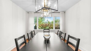 Ein Esszimmer mit einem langen Tisch, Stühlen, einer Kristallleuchter und großen Fenstern mit Blick auf Grünflächen.