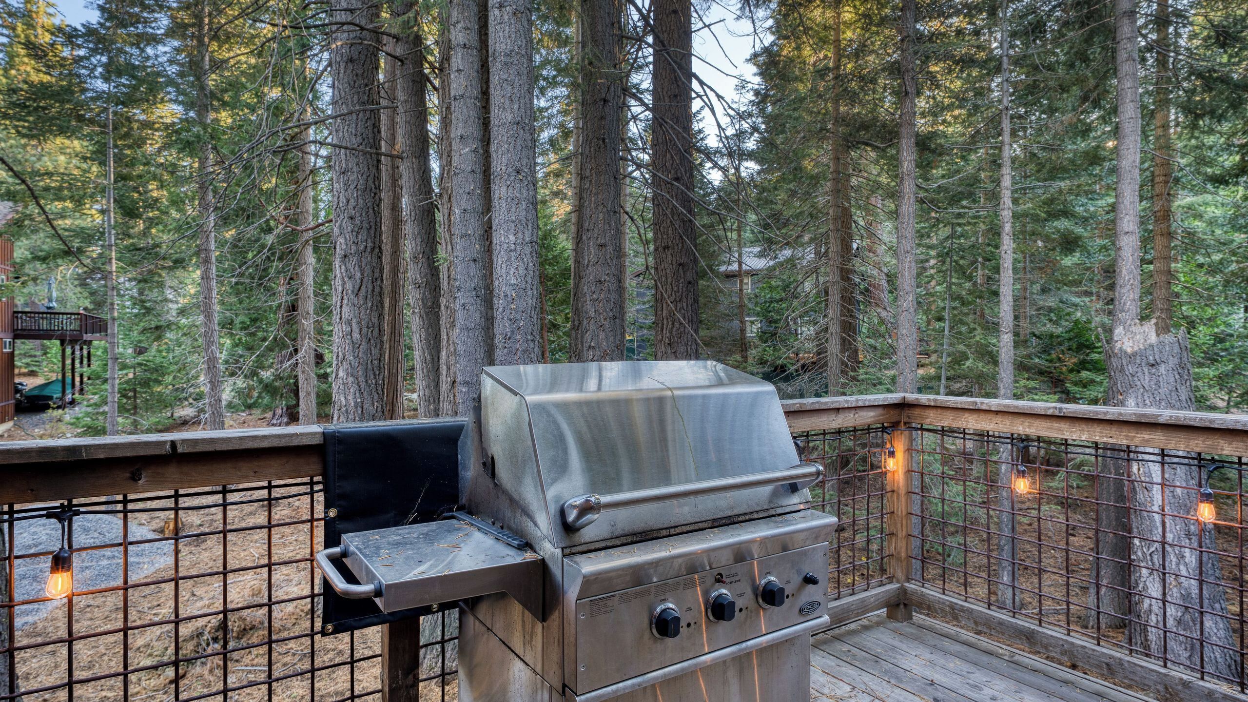 A metal grill on an outdoor deck with a wooden railing, surrounded by tall trees.