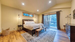 A bedroom with a large bed, a wooden bench, a dresser, a wicker basket, and a view of the outdoors through a large window. - Cozy Tahoe Retreat w/ Hot Tub, Movie Room & Forest Views 10th ave 2 by Avantstay (Tahoma)