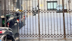 En grind med ett staket av metall, en parkerad skoter innanför grinden och en gatuvy med byggnader i bakgrunden.