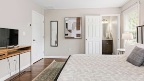 A bedroom with a bed, a TV on a wooden stand, a mirror, and a bathroom visible through an open door.