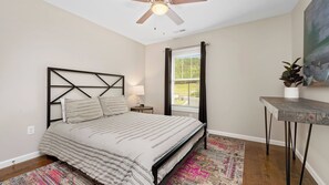 A bedroom with a bed, a ceiling fan, a window with curtains, a small table with a plant, and a colorful rug.
