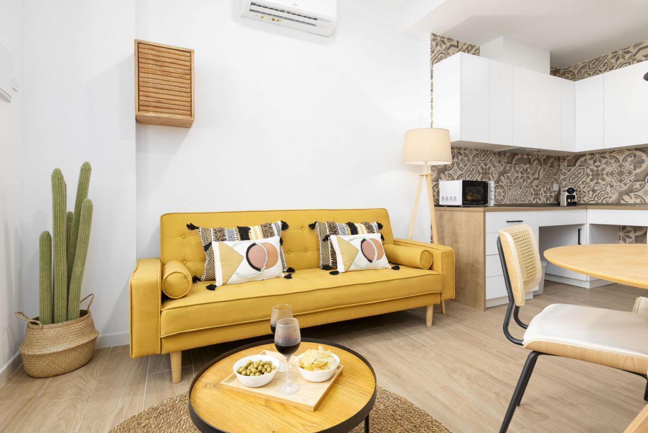 A yellow sofa with patterned cushions, a round wooden table with bowls and glasses, a cactus plant, and a kitchen area with white cabinets and a microwave.