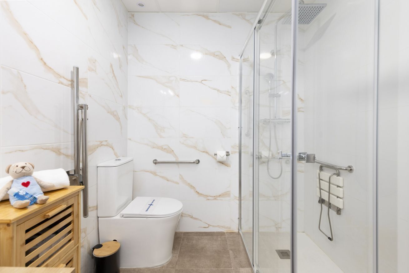 A bathroom with a marble wall, a glass shower enclosure, a toilet, and a wooden vanity with a teddy bear.