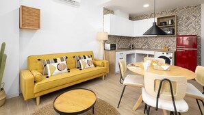 Un espace de vie compact comprenant un canapé jaune, une table ronde en bois et un coin cuisine équipé d’un four à micro-ondes, d’un grille-pain et d’un réfrigérateur rouge.