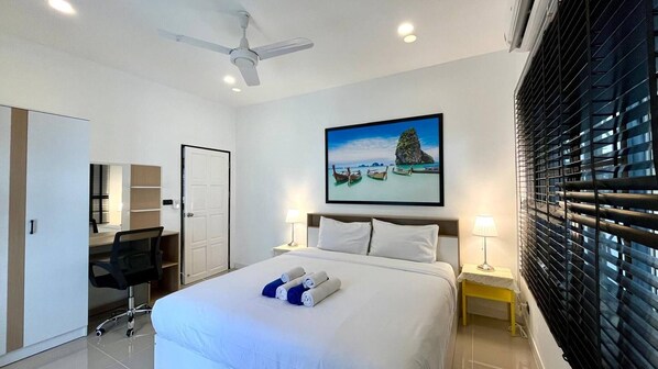 Une chambre d’hôtel avec un lit, un bureau, une chaise, un ventilateur de plafond, une fenêtre avec des stores et un tableau au mur.