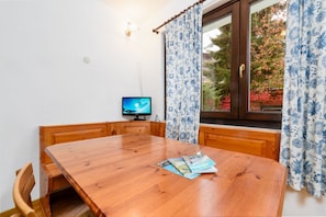A wooden dining table with a magazine, a television on a stand, and a window with curtains.