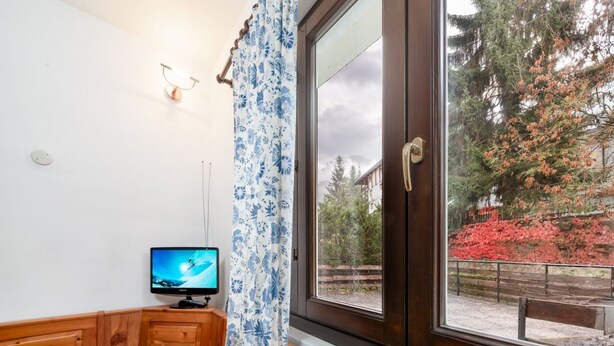 A room with a wooden bench, a television on a stand, a window with a curtain, and a view of trees and a fence outside. - Casa Daisy 2 by Rentbeat  (Limone)