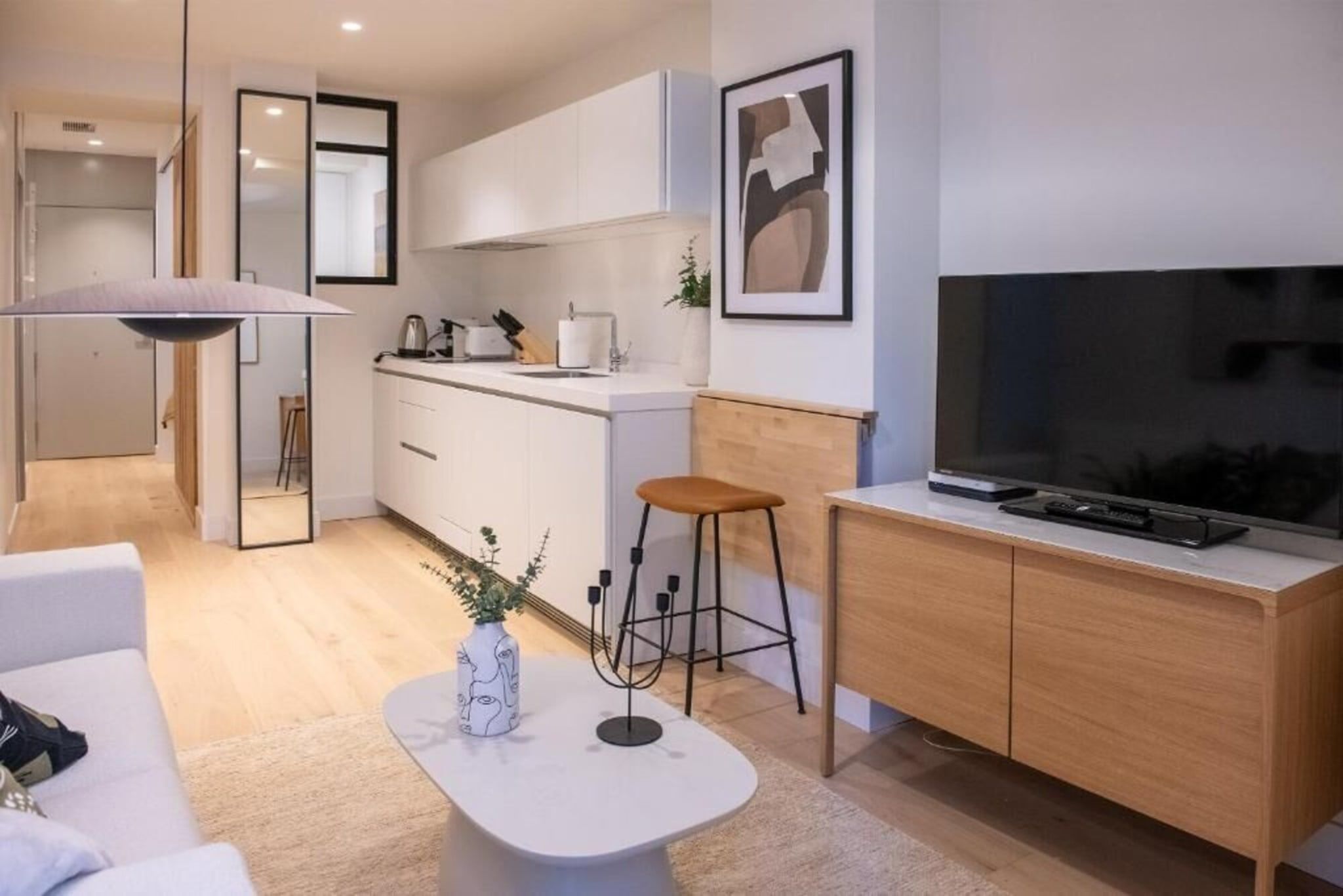 Een moderne woonkamer met een witte bank, een salontafel met een vaas, een televisie op een houten kast en een keuken op de achtergrond.