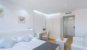 Une chambre d’hôtel moderne équipée d’un lit, d’une table de chevet, d’une chaise, d’un bureau et d’une petite table sur laquelle sont disposés des fruits.