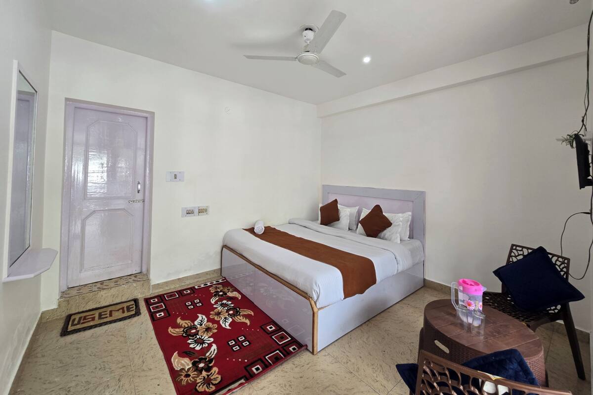 Une chambre à coucher avec un lit, un ventilateur de plafond, une petite table sur laquelle se trouve une bouteille d’eau rose, et une porte.