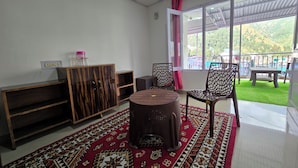 A room with a patterned rug, a round table, two chairs, a cabinet, and a large window with a view of greenery. - Triund View Homestay (Dharamshala)