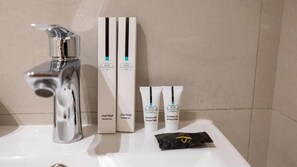 A bathroom sink with a chrome faucet, two boxes labeled "Acacia Dental Kit," two tubes of "Acacia Shower Gel," and a black packet with a gold emblem.