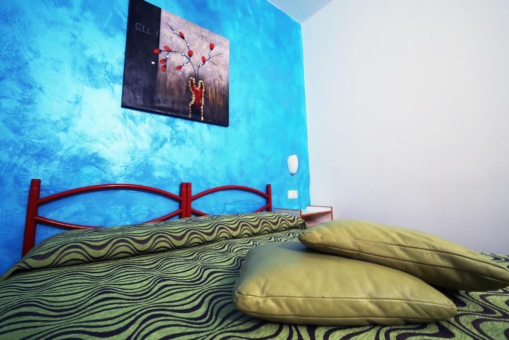 Une chambre avec un mur bleu à texture, un cadre de lit en métal rouge, une literie à motifs et une œuvre d’art accrochée au mur.