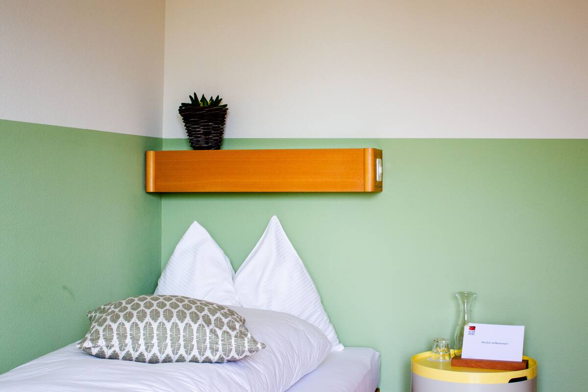 Una cama bien hecha con almohadas blancas y estampadas, una planta pequeña en un estante superior y una mesita de noche con un florero y una tarjeta.