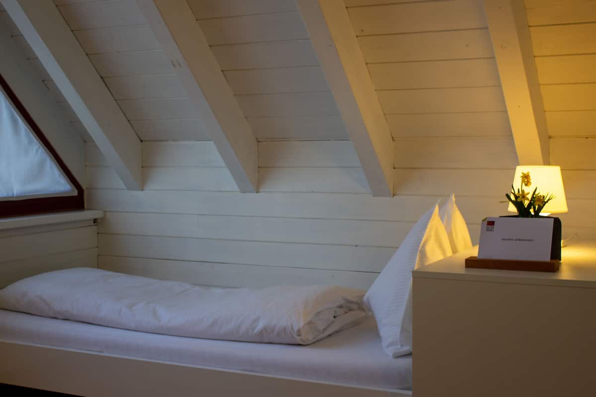 Una cama individual en una habitación con techos inclinados y paneles de madera. Se ve una mesita de noche con una lámpara y una tarjeta de bienvenida.