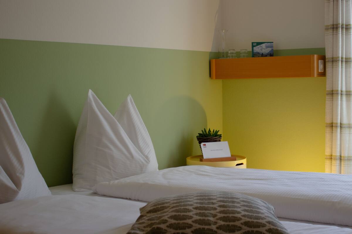 Habitación de hotel con dos camas, una mesita auxiliar, pared verde y amarilla, y una ventana con cortinas.
