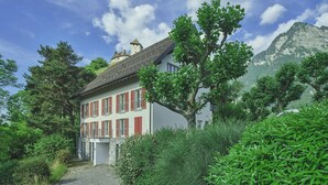 Une maison à deux étages avec des volets rouges, entourée d’une végétation luxuriante et dominée par une chaîne de montagnes en arrière-plan.