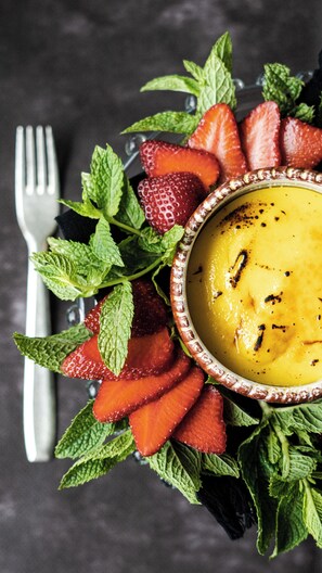 A bowl of yellow soup garnished with black pepper, surrounded by strawberries and mint leaves. - Rouge Maison (Milano Marittima)