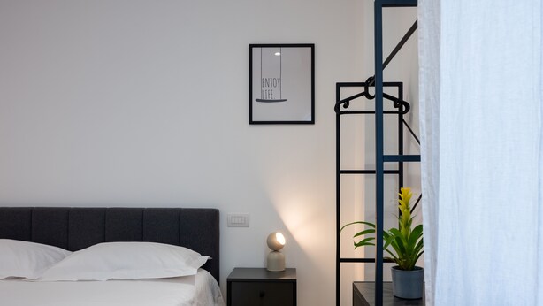 A neatly made bed with white linens, a bedside table with a lamp, a framed picture on the wall, and a potted plant. - Cosy is a very cozy modern facility in soft colors, center of Bari (Bari)