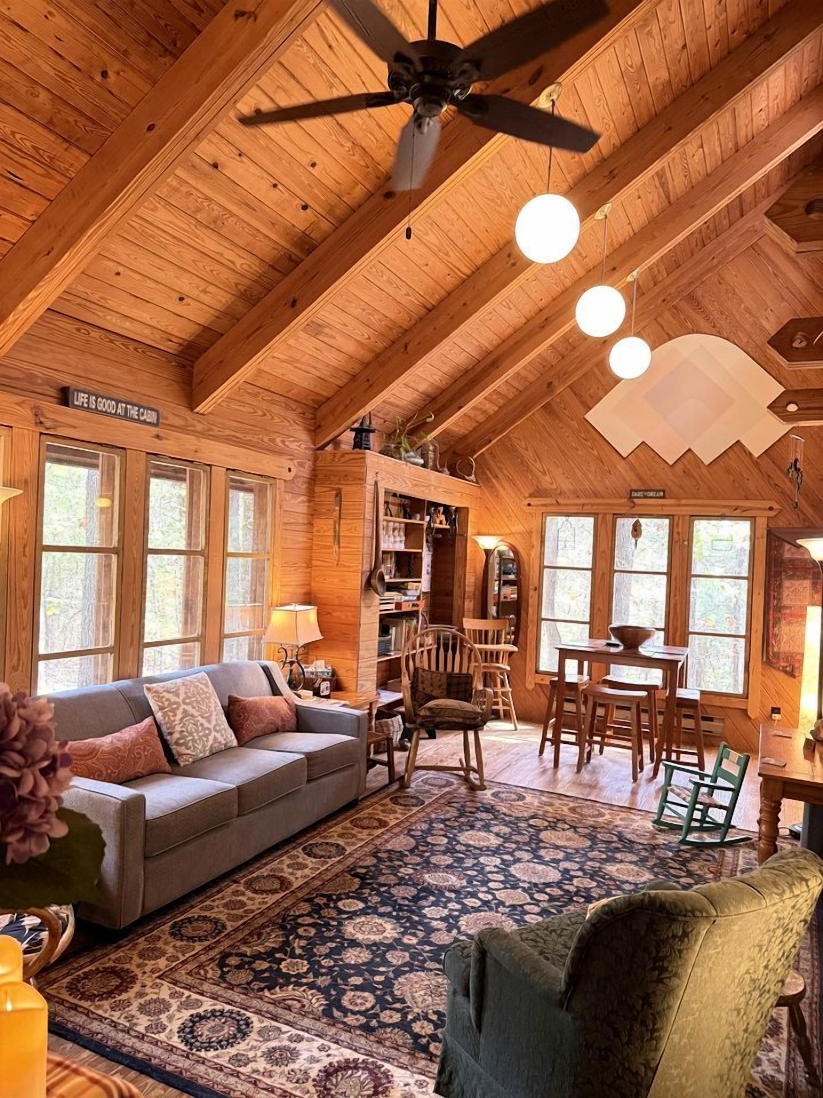 A cozy cabin interior with a wooden ceiling, a sofa, a dining table, and a ceiling fan.