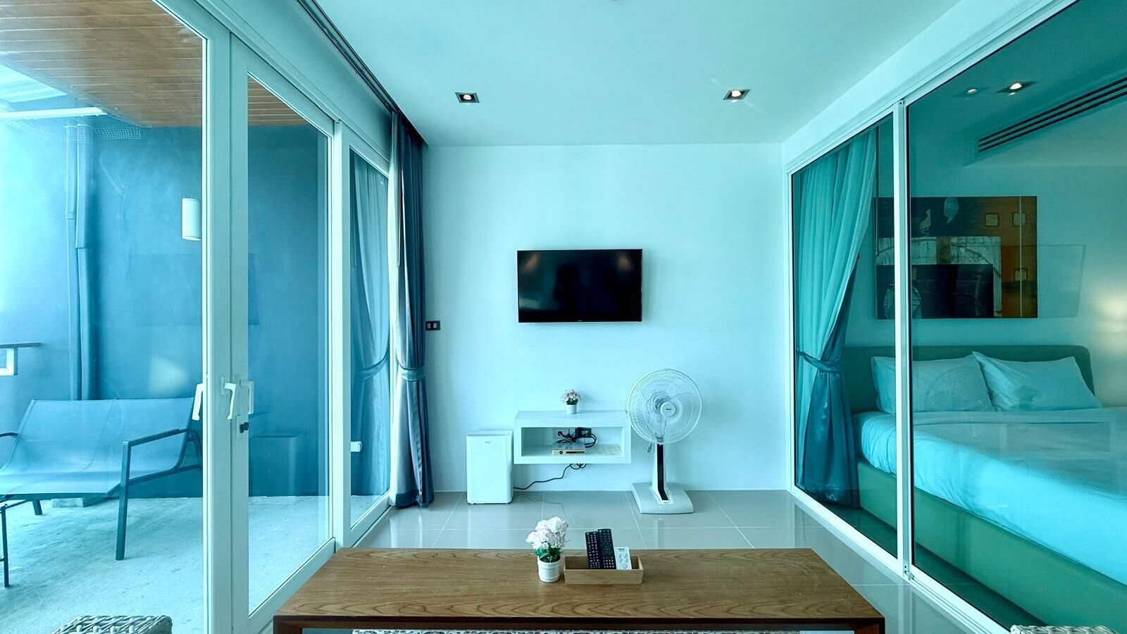 A modern hotel room with a large bed, a TV mounted on the wall, a small white cabinet, a fan, and a wooden coffee table with a vase and remote controls.