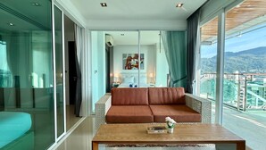 A modern hotel room with a large balcony, a brown sofa, a wooden coffee table, and a city view. - Emerald Terrace Patong (Patong)