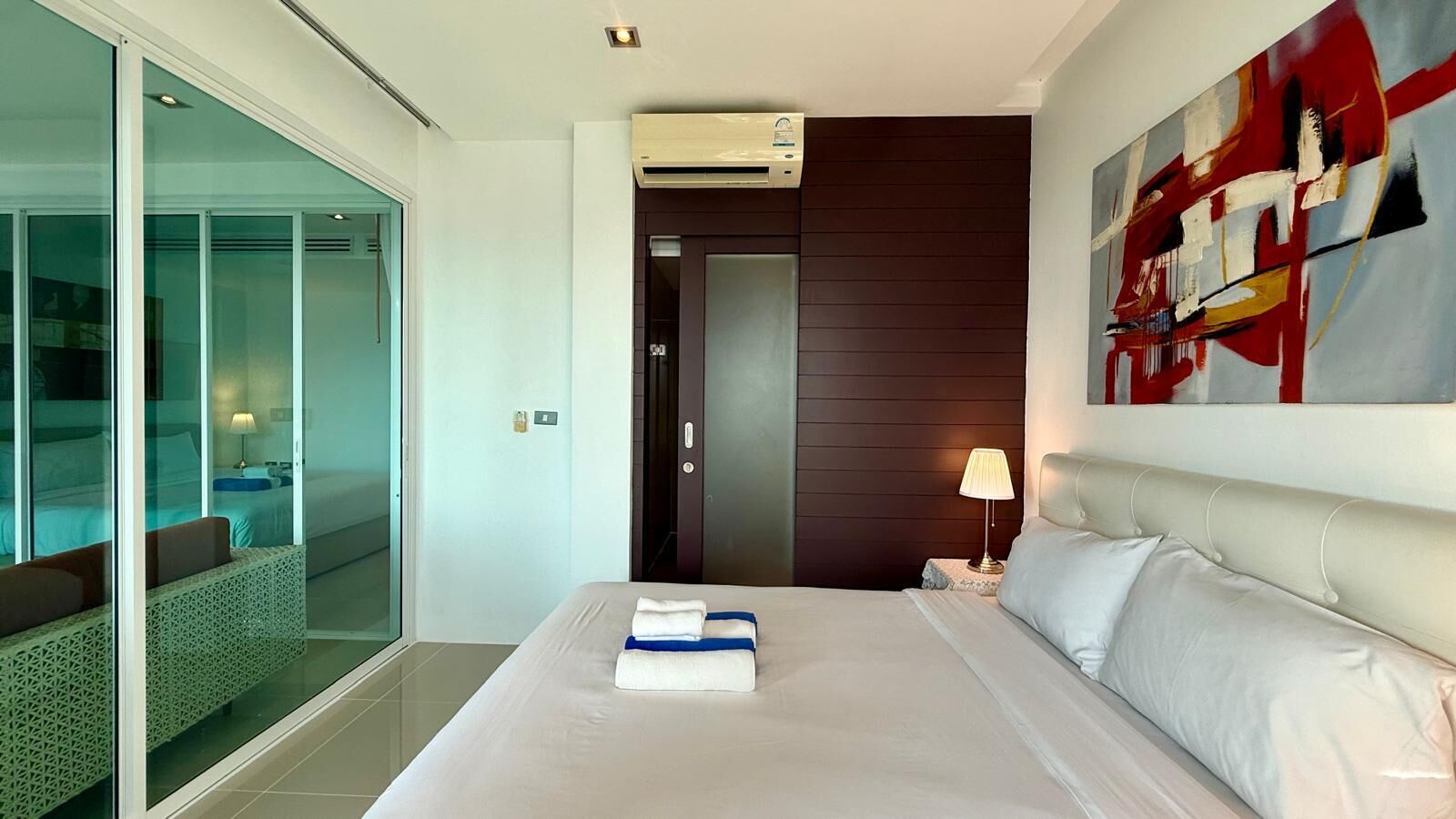 A hotel room with a bed, a glass door leading to a balcony, a painting on the wall, and an air conditioning unit.