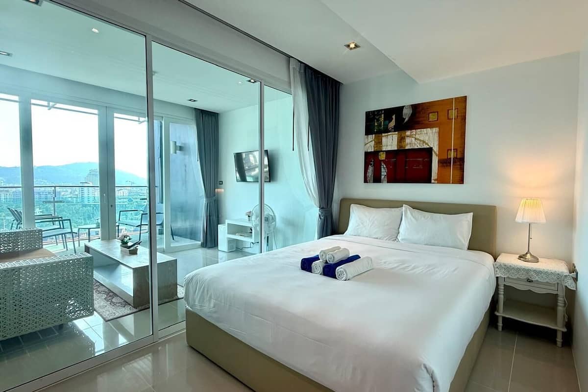 A modern hotel room with a large bed, a glass door leading to a balcony, a flat-screen TV, and a view of the city.