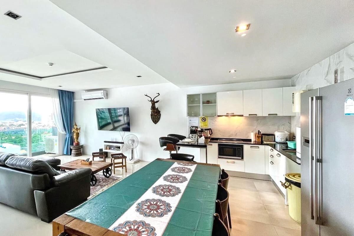 A modern living room with a sofa, a dining table with a patterned runner, a flat-screen TV, and a kitchen area with cabinets and appliances.