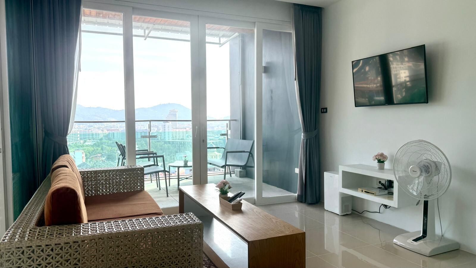 A modern living room with a sofa, a coffee table, a TV mounted on the wall, and a balcony with a view of the city.