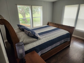 A bedroom with a wooden bed, a nightstand, and a lamp. There are windows with blinds, and a view of greenery outside.
