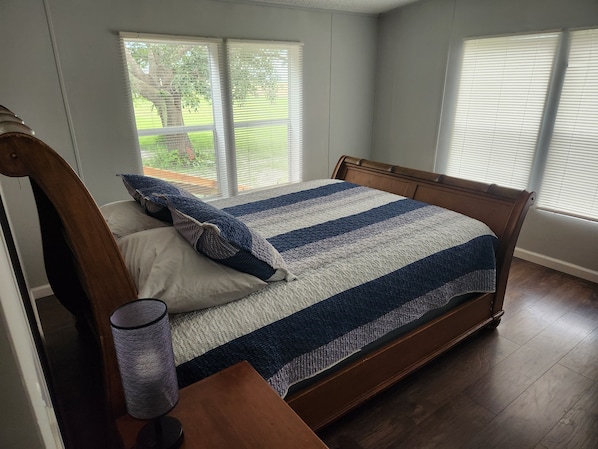 A bedroom with a wooden bed, a nightstand, and a lamp. There are windows with blinds, and a view of greenery outside.