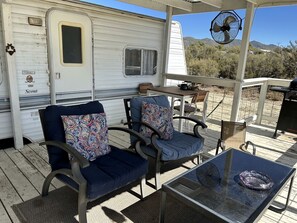 A covered outdoor seating area with two chairs and a table, a white RV in the background, and a fan on the roof.