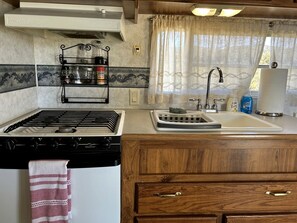 A compact kitchen with a gas stove, a sink, wooden cabinets, and a window with curtains. - Shabby Chic Warner Springs Rental at a Ranch Ideal for Glamping in California (Warner Springs)