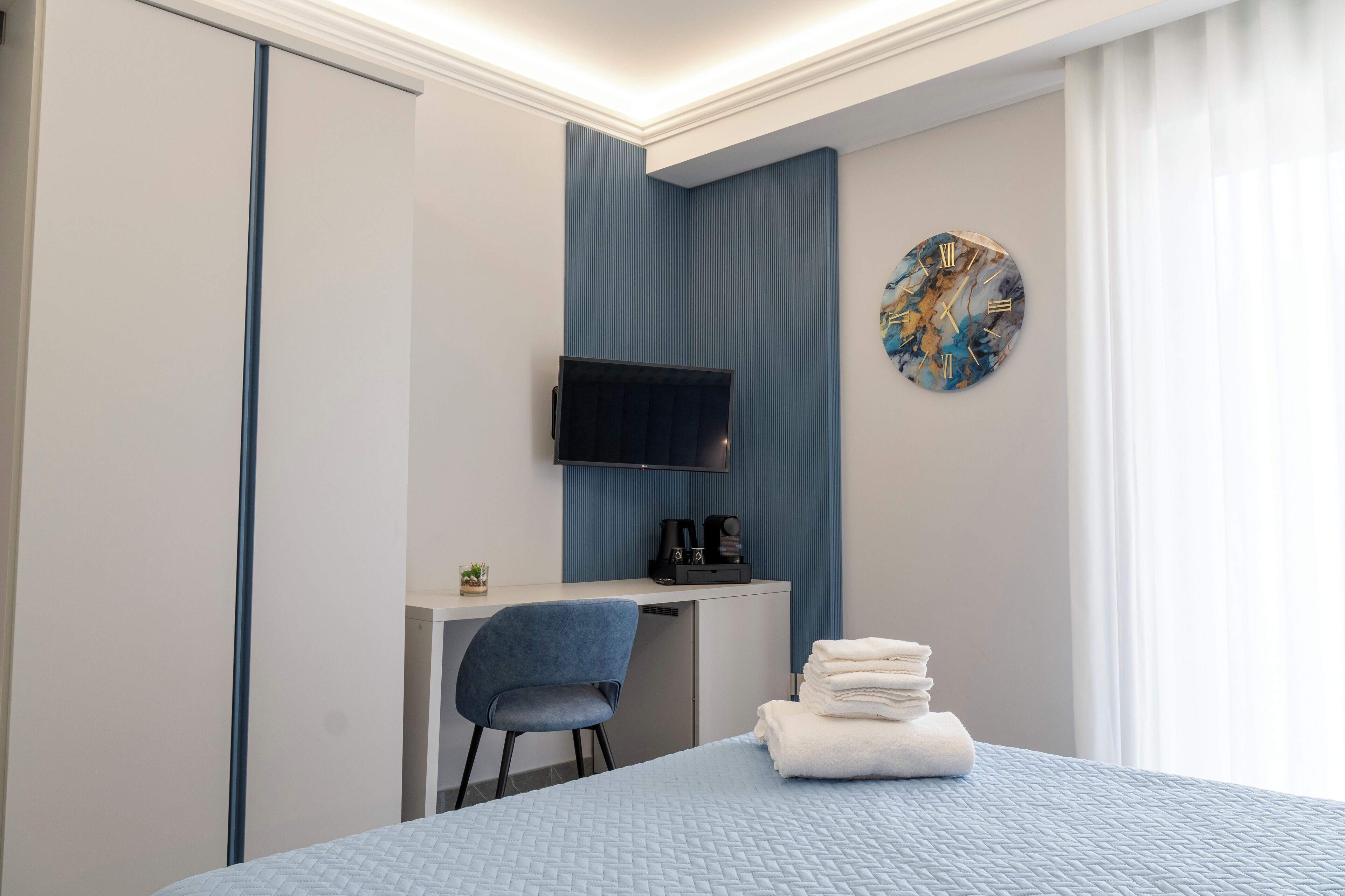 Ein Hotelzimmer mit einem Bett, einem Schreibtisch mit Stuhl, einem an der Wand befestigten Flachbildfernseher und einer Uhr.