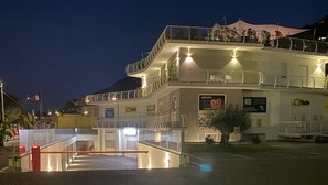 Un edificio de varios pisos con un nivel superior cubierto, una escalera que lleva a la entrada y un área pavimentada al frente.