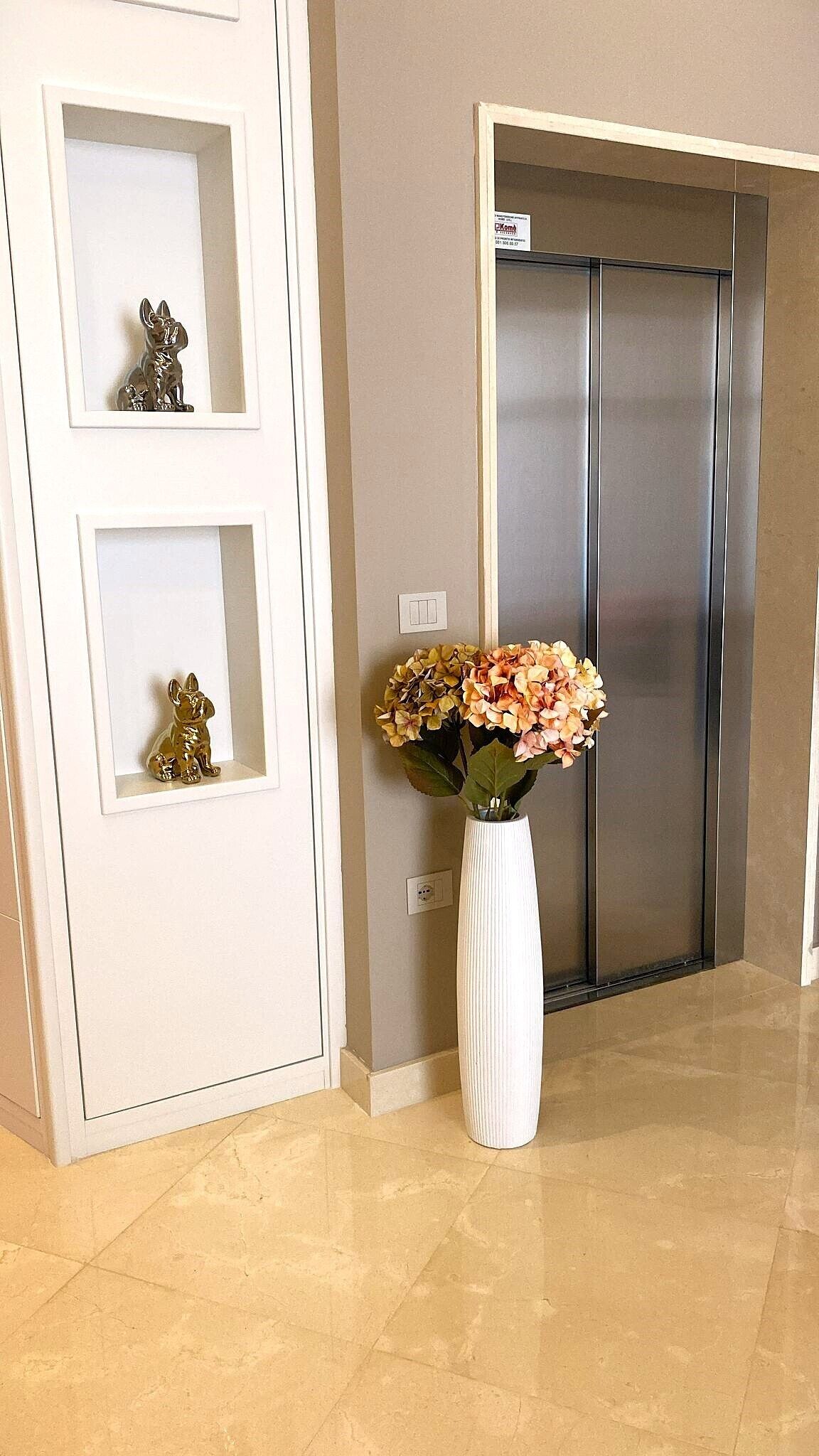 A hallway with a white door, a white vase with flowers, and a silver elevator.