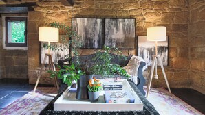 Een gezellige woonkamer met een stenen muur, een bank, twee vloerlampen, een bijzettafeltje met boeken en een plant, en een raam met uitzicht op groen.