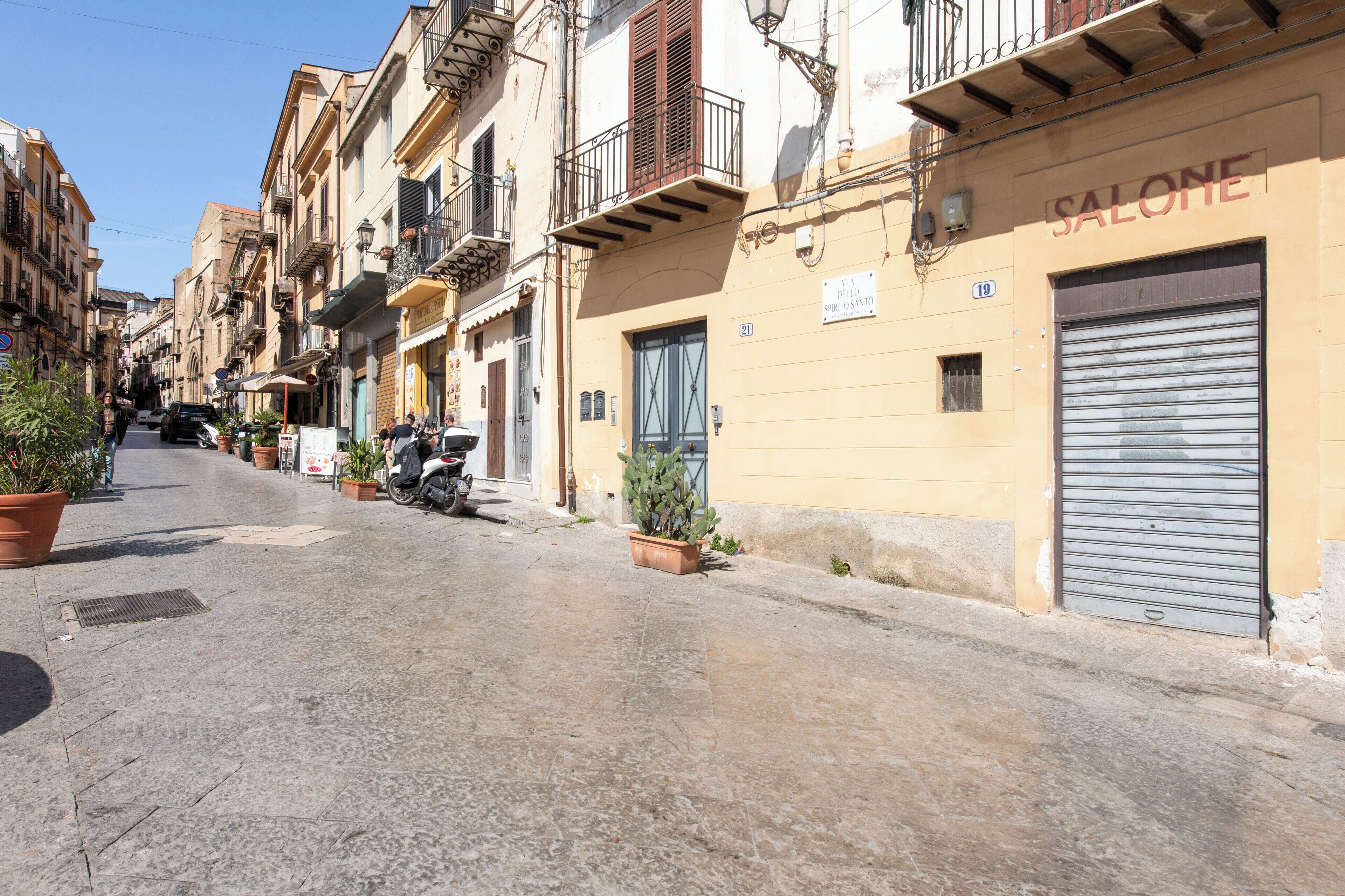 A narrow street lined with multi-story buildings, some with balconies and potted plants.