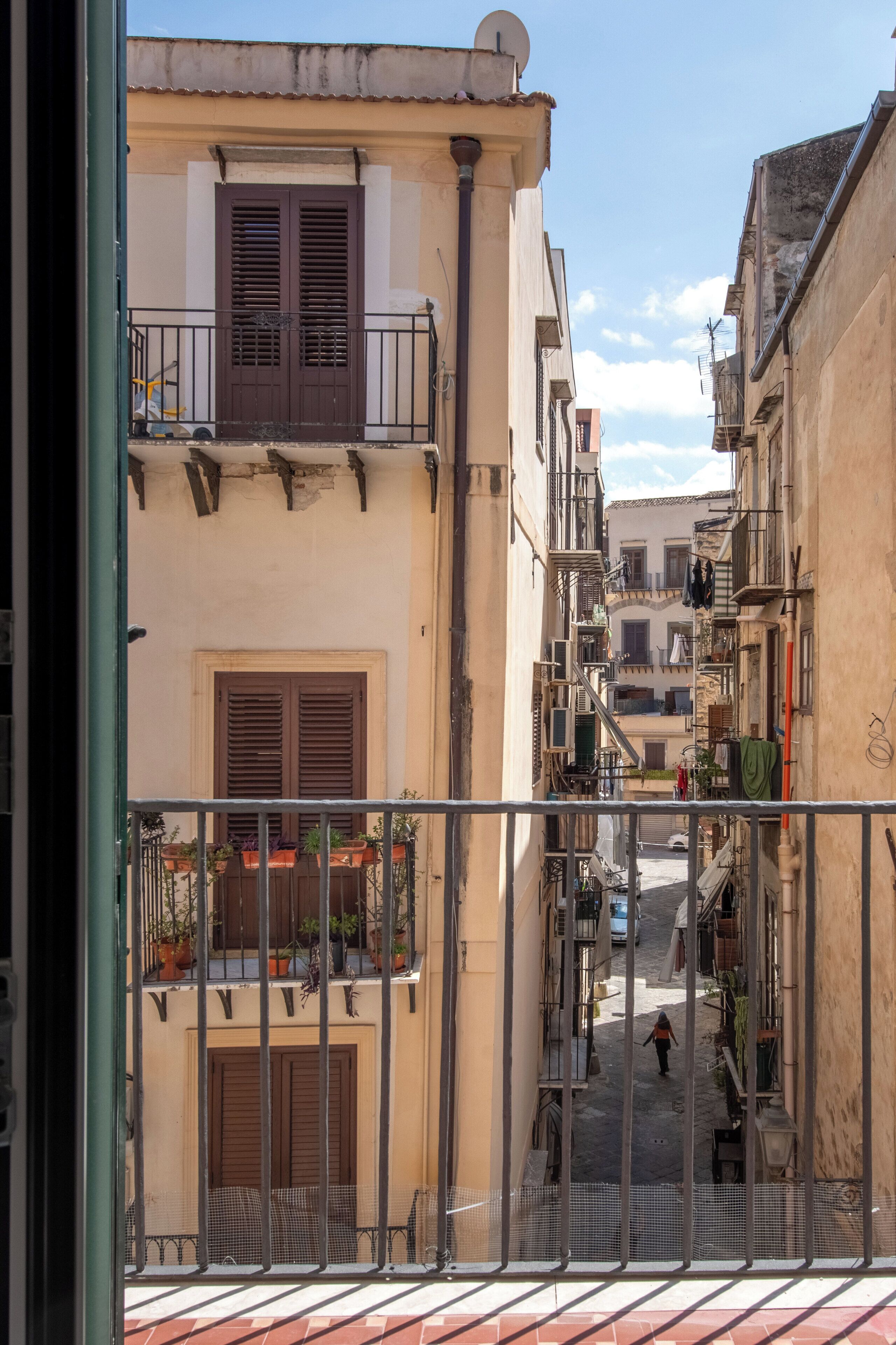 Un carrer estret amb edificis a ambdós costats, balcons amb plantes en testos, i una persona caminant pel carrer.