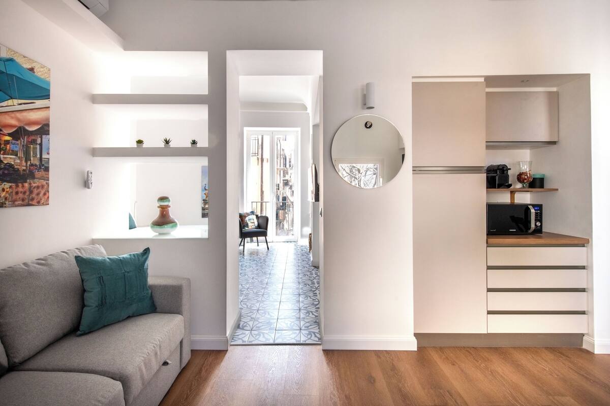 Un appartement moderne avec un canapé gris, une cuisine équipée d’un four à micro-ondes et un couloir menant à un espace ouvert.