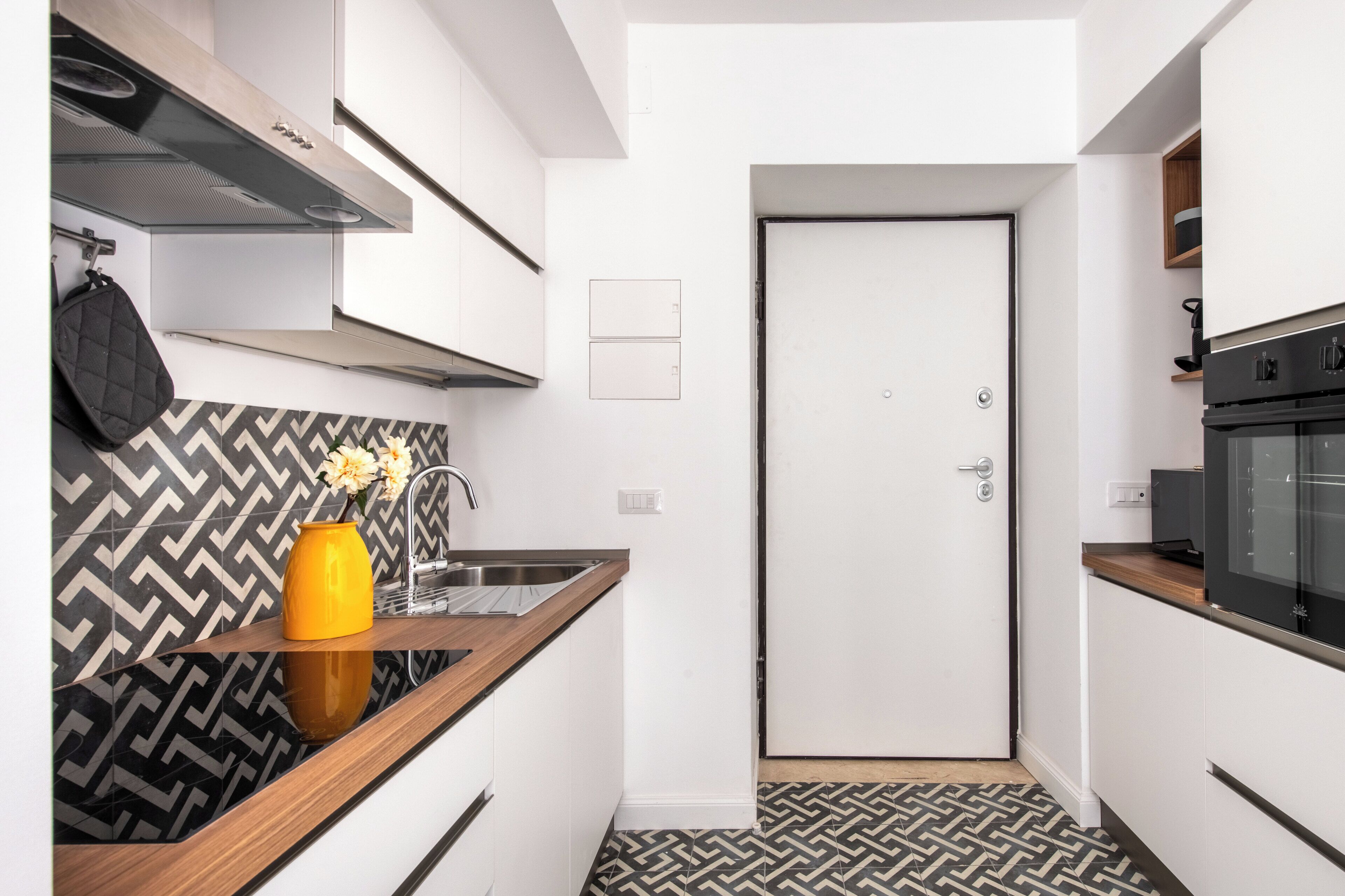Une cuisine compacte avec des meubles blancs, un crédence à motifs noir et blanc, un plan de travail en bois et une porte dotée d’une poignée.
