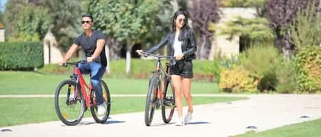 Dos personas en bicicleta sobre un camino pavimentado, con vegetación de fondo.