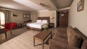 Habitación de hotel con cama, una silla roja, un sofá marrón, una mesa de centro de madera y una mesita con un florero.