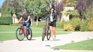Två personer cyklar på en asfalterad stig i en park, med grönska och stenstrukturer i bakgrunden.